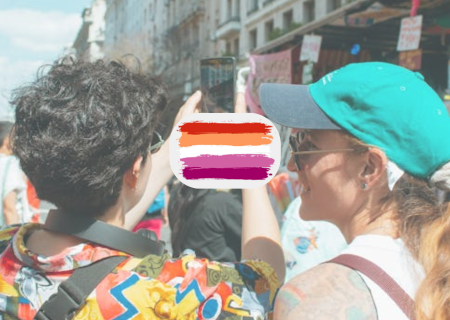 Dues noies al carrer. Un d'elles treu una foto amb mòbil. Entre elles, la bandera lèsbica